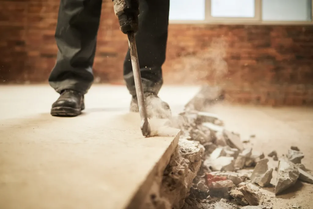 Sloopwerk: gebroken dekvloer-brokken op stapel, houten balklaag zichtbaar, arbeider met bezem op de achtergrond